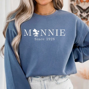 K&ouml;nnte beinhalten: Ein blaues Sweatshirt mit wei&szlig;em Text, der "M*NNIE Since 1928" lautet. Der Text ist mittig auf dem Sweatshirt platziert und zeigt eine kleine Grafik einer Maus.