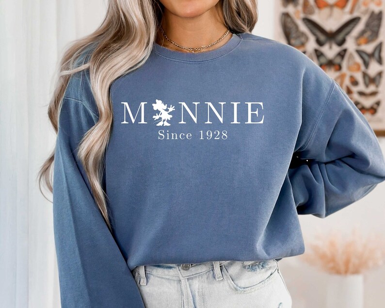 K&ouml;nnte beinhalten: Ein blaues Sweatshirt mit wei&szlig;em Text, der "M*NNIE Since 1928" lautet. Der Text ist mittig auf dem Sweatshirt platziert und zeigt eine kleine Grafik einer Maus.