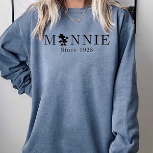 K&ouml;nnte beinhalten: Ein blaues Sweatshirt mit einer schwarzen Grafik einer Comic-Maus und dem Text "MNNIE Since 1928"