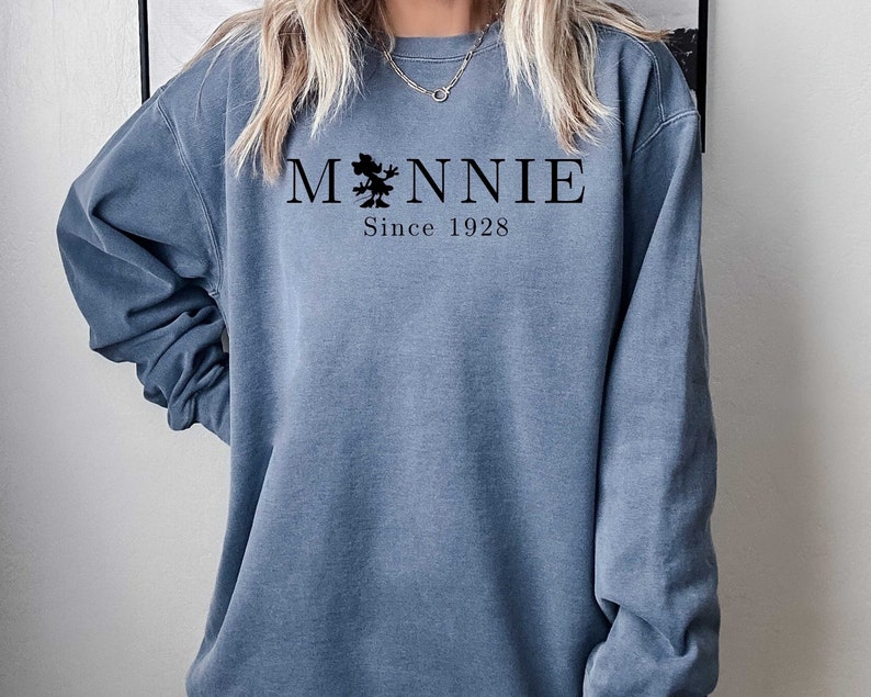 K&ouml;nnte beinhalten: Ein blaues Sweatshirt mit einer schwarzen Grafik einer Comic-Maus und dem Text "MNNIE Since 1928"