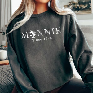 K&ouml;nnte beinhalten: Ein dunkelgrauer Pullover mit einem wei&szlig;en Grafikdesign, auf dem "M*NNIE Since 1928" steht.