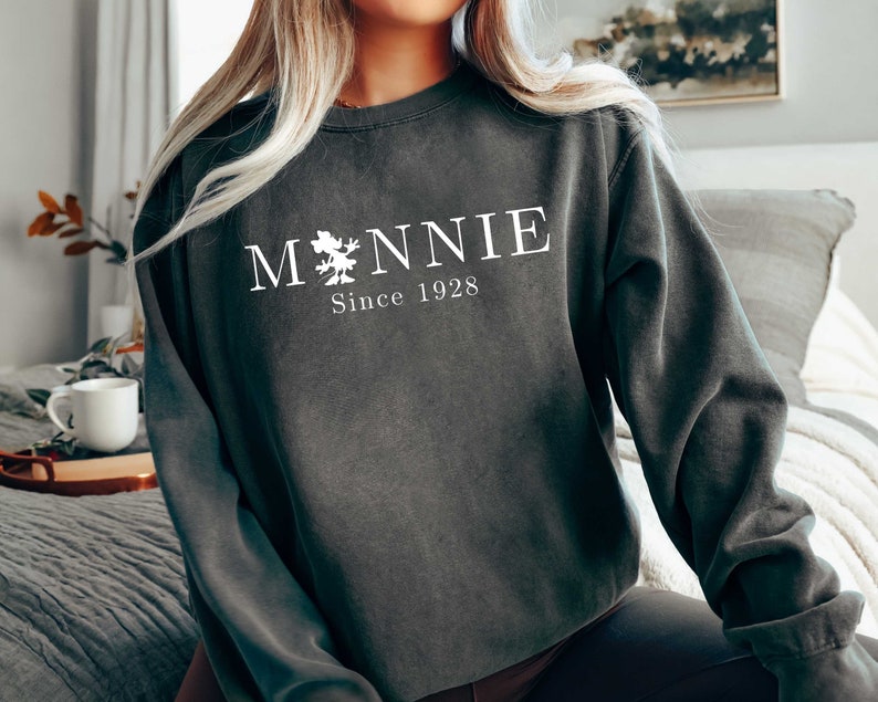 K&ouml;nnte beinhalten: Ein dunkelgrauer Pullover mit einem wei&szlig;en Grafikdesign, auf dem "M*NNIE Since 1928" steht.