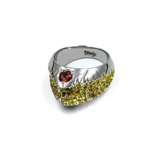 May include: A silver and gold ring with a red gemstone set in the center. The ring has a textured design and is marked "Deutsch Jewelry".