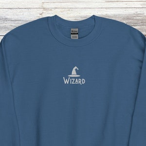 May include: A teal sweatshirt featuring the word "Wizard" and a wizard hat icon embroidered in white. The crew neck sweatshirt is a solid colour. The background includes dice and a map.