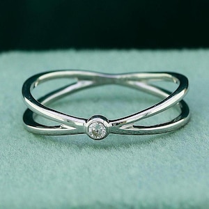 Può includere: Un anello d'argento con una singola piccola pietra preziosa trasparente incastonata al centro. L'anello ha un design semplice ed elegante con due bande che si incrociano.