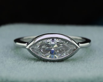May include: A silver ring with a marquise-shaped diamond set in a bezel setting. The ring is on a light green velvet cushion.