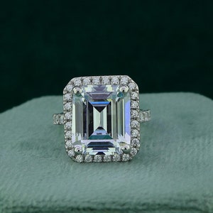 May include: A silver ring with a large, clear emerald-cut gemstone surrounded by a halo of smaller diamonds. The ring is displayed on a green velvet box.
