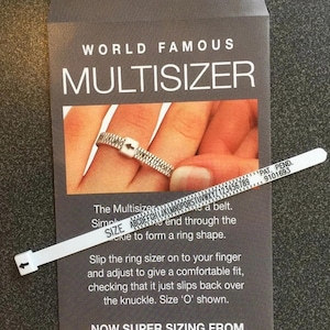May include: A white plastic ring sizer tool with a buckle and printed alphabet and numeric scale, presented on a dark grey instructional card labelled WORLD FAMOUS MULTISIZER. The card features text instructions and an image of the tool in use.