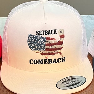 Könnte beinhalten: Weiße Trucker-Kappe mit einer gestickten amerikanischen Flagge in Rot, Weiß und Blau mit dem Text "Setback Comeback" und einem Etikett "The Authentic Yupoong Snapback Since 1974".