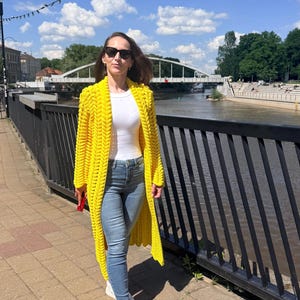 May include: A bright yellow, long-length, chunky knit cardigan featuring a prominent braided stitch pattern, worn over a plain white short-sleeved top and light blue denim jeans with white sneakers and dark-tinted sunglasses.