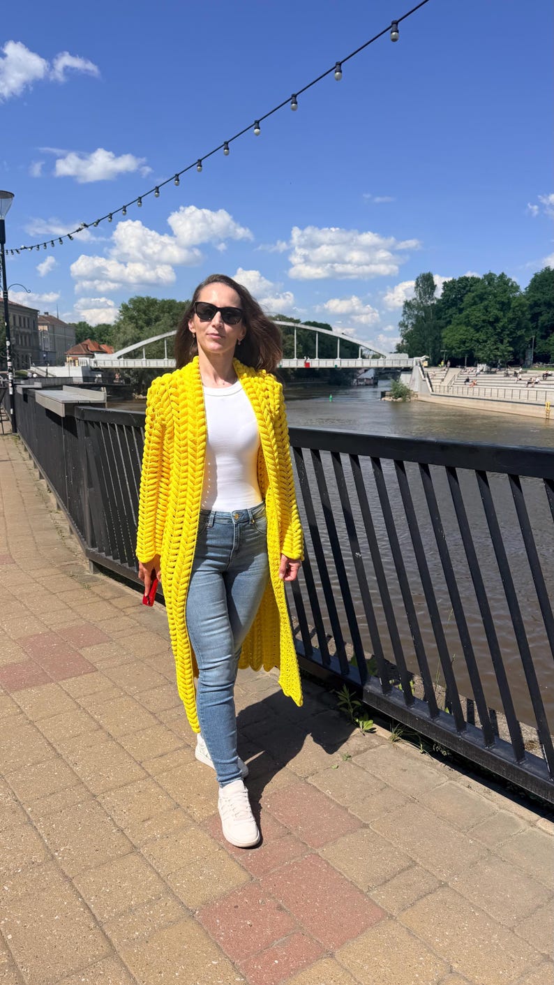 May include: A bright yellow, long-length, chunky knit cardigan featuring a prominent braided stitch pattern, worn over a plain white short-sleeved top and light blue denim jeans with white sneakers and dark-tinted sunglasses.