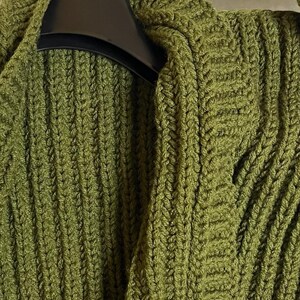 May include: Close-up of a green knitted cardigan with a textured, ribbed design. The cardigan is hanging on a black hanger. The olive green color and thick knit suggest warmth and comfort, perfect for cooler weather.
