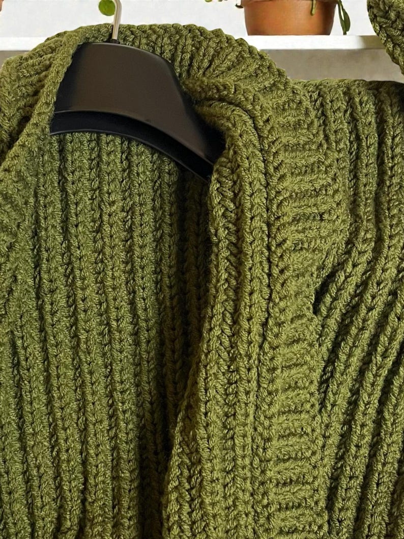 May include: Close-up of a green knitted cardigan with a textured, ribbed design. The cardigan is hanging on a black hanger. The olive green color and thick knit suggest warmth and comfort, perfect for cooler weather.