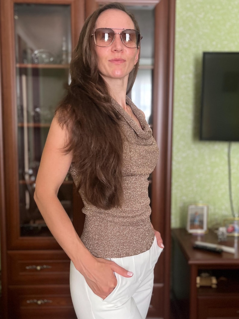 May include: A sleeveless, metallic gold top with a cowl neck is paired with white pants. The person is wearing large, square-framed sunglasses with a brown tint. The outfit is suitable for a casual or semi-formal occasion.