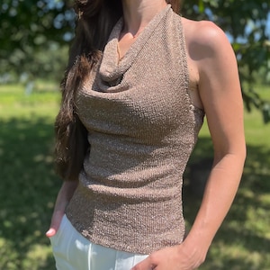 May include: A metallic gold halter top with a cowl neck. The top is sleeveless and has a ribbed texture. The model is wearing white pants. The background is a blurred green outdoor scene.
