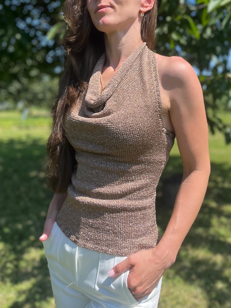 May include: A metallic gold halter top with a cowl neck. The top is sleeveless and has a ribbed texture. The model is wearing white pants. The background is a blurred green outdoor scene.