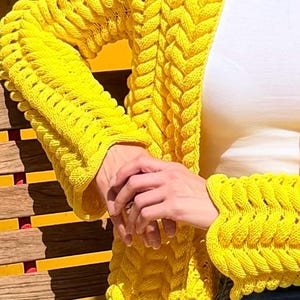 May include: A bright yellow open-front cardigan featuring a prominent, chunky braided knit pattern that runs vertically along the front panels and down the length of the long sleeves, ending in wide, textured cuffs with a distinct ribbed edge.