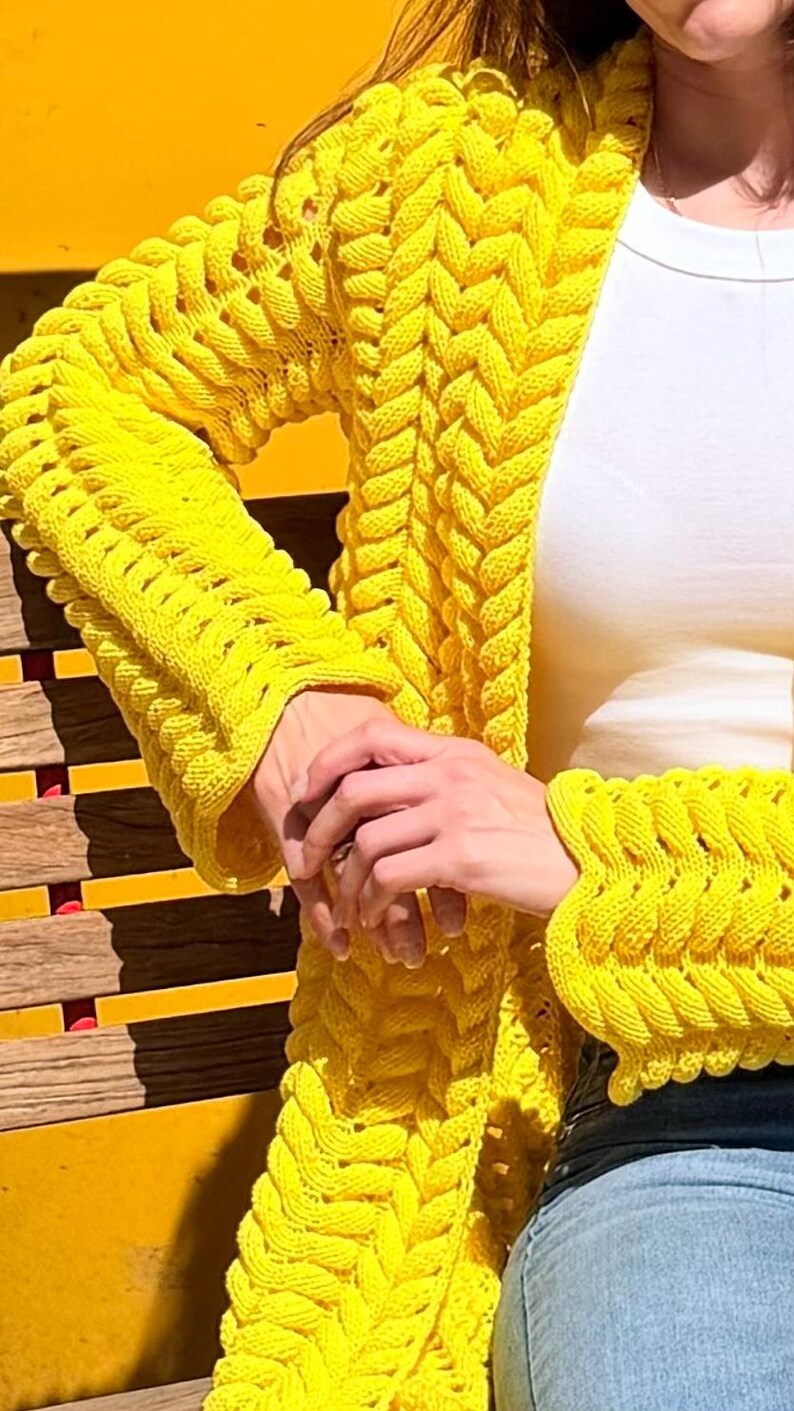 May include: A bright yellow open-front cardigan featuring a prominent, chunky braided knit pattern that runs vertically along the front panels and down the length of the long sleeves, ending in wide, textured cuffs with a distinct ribbed edge.