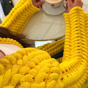 May include: A bright yellow long-sleeved sweater featuring a chunky, textured knit pattern with raised, braided-style vertical columns and openwork detailing. The garment has a round neckline and a thick, prominent knit construction throughout.