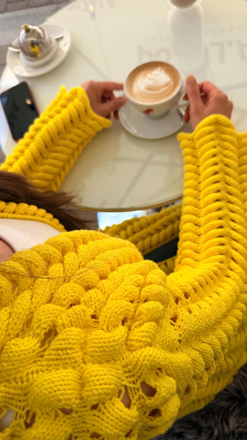 May include: A bright yellow long-sleeved sweater featuring a chunky, textured knit pattern with raised, braided-style vertical columns and openwork detailing. The garment has a round neckline and a thick, prominent knit construction throughout.