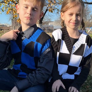 Peut inclure: Deux enfants portant des gilets sans manches à carreaux. L'un est bleu et noir, l'autre est noir et blanc. Les deux gilets ont une encolure en V. Les enfants sont assis dehors par une journée ensoleillée.