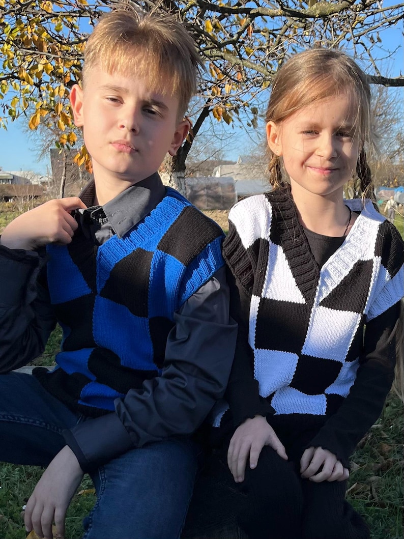 May include: Two children wearing checkered sweater vests. One vest is blue and black, the other is black and white. Both vests have a V-neck design. The children are sitting outside on a sunny day.