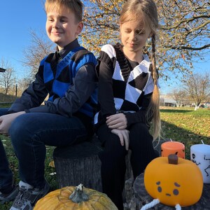 May include: Two children sit on tree stumps, wearing checkered vests. One vest is blue and black, the other black and white. A carved pumpkin with a face and legs sits in front of them, along with candles. A large pumpkin is on the left.