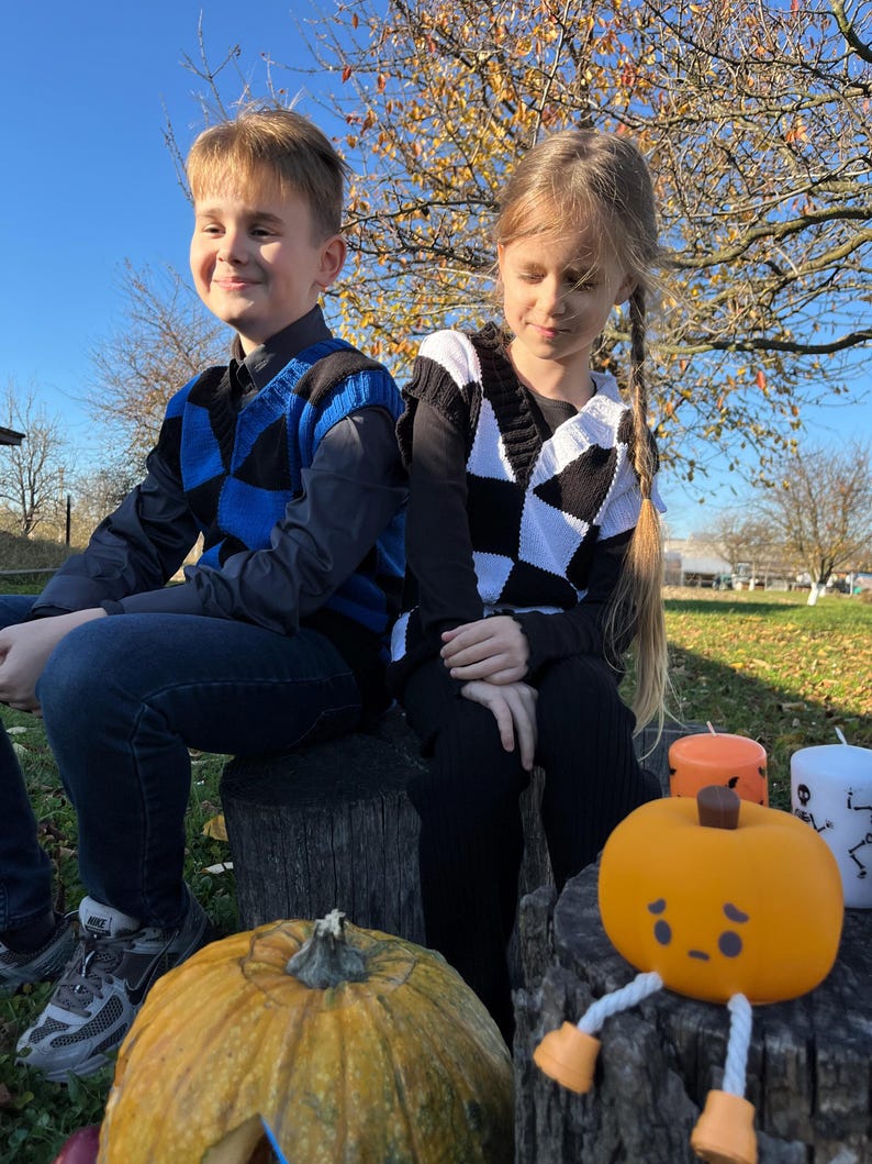 May include: Two children sit on tree stumps, wearing checkered vests. One vest is blue and black, the other black and white. A carved pumpkin with a face and legs sits in front of them, along with candles. A large pumpkin is on the left.