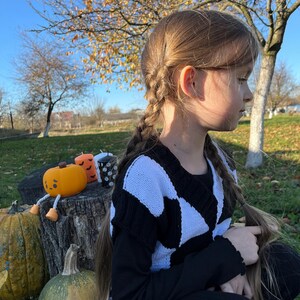 May include: A young person with long braided hair wearing a black and white patterned sweater. The scene includes pumpkins and decorative candles on a tree stump, suggesting a Halloween theme. The background features trees and a clear sky.