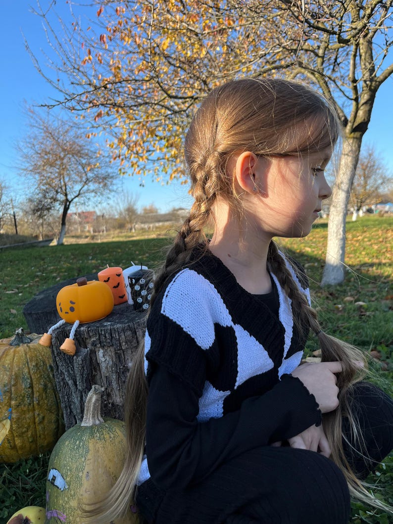 May include: A young person with long braided hair wearing a black and white patterned sweater. The scene includes pumpkins and decorative candles on a tree stump, suggesting a Halloween theme. The background features trees and a clear sky.
