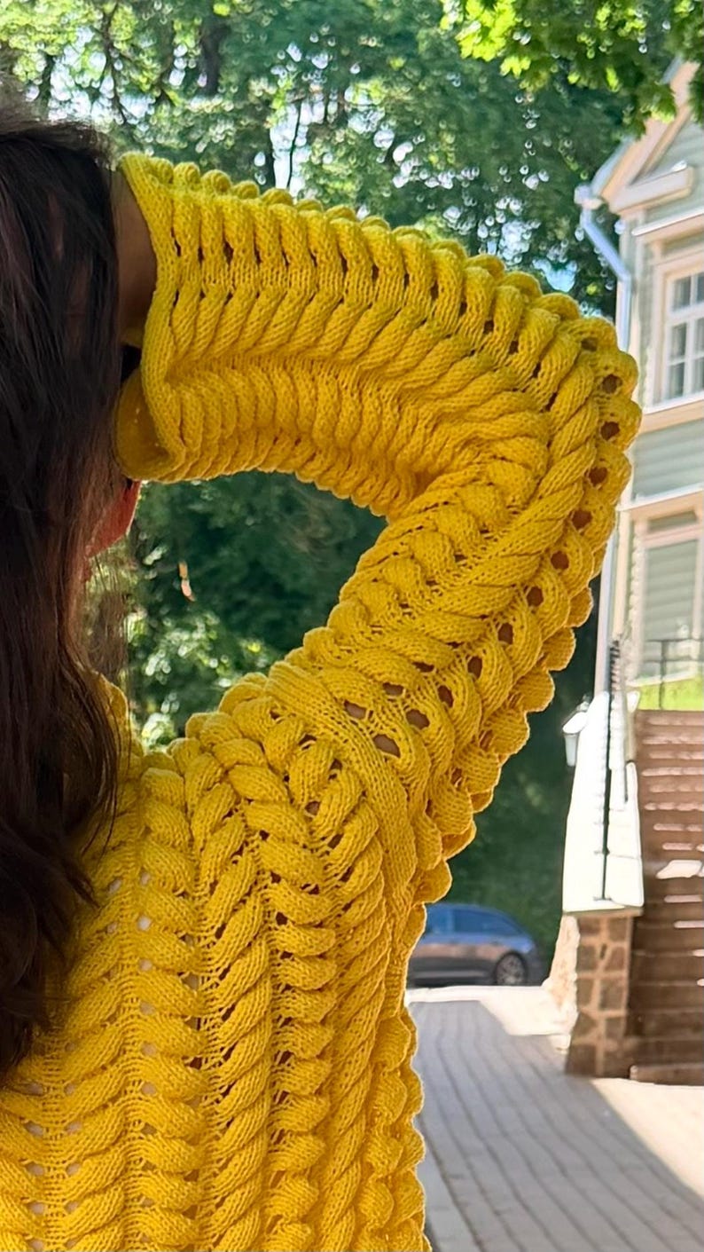 May include: A bright yellow long-sleeve sweater featuring a prominent vertical cable knit pattern with openwork detailing between the textured columns. The garment has a high neckline and a thick, chunky knit construction throughout the sleeves and body.
