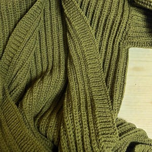 May include: Olive green knitted cardigan with a shawl collar. The sweater is made of a thick, textured knit fabric. The cardigan is displayed on a wooden surface.