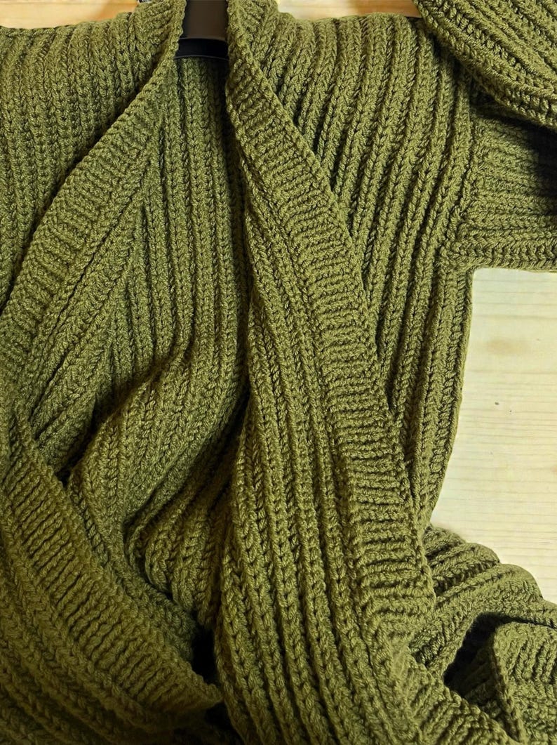 May include: Olive green knitted cardigan with a shawl collar. The sweater is made of a thick, textured knit fabric. The cardigan is displayed on a wooden surface.