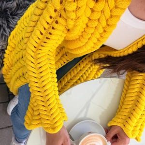 May include: A bright yellow chunky knit cardigan featuring a prominent braided cable stitch pattern, long sleeves, and an open front design. The garment is worn over a white top and blue denim jeans, paired with white sneakers.