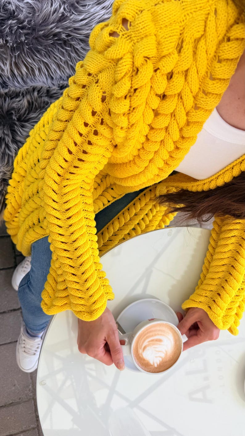 May include: A bright yellow chunky knit cardigan featuring a prominent braided cable stitch pattern, long sleeves, and an open front design. The garment is worn over a white top and blue denim jeans, paired with white sneakers.