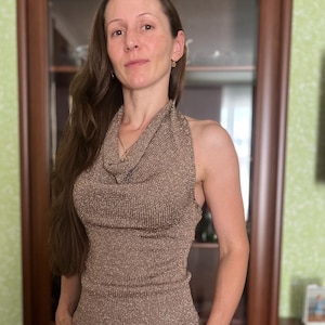 May include: A sleeveless, metallic brown top with a cowl neck. The top is paired with white pants. The woman in the image is wearing a gold necklace and earrings. The background includes a wooden cabinet and a green wall.