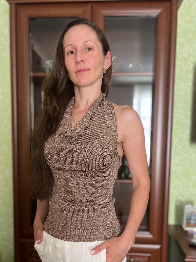 May include: A sleeveless, metallic brown top with a cowl neck. The top is paired with white pants. The woman in the image is wearing a gold necklace and earrings. The background includes a wooden cabinet and a green wall.