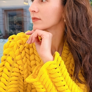 May include: A bright yellow long-sleeved sweater featuring a chunky, braided cable knit pattern with a V-neckline. The garment is constructed with thick, textured yarn, showcasing a prominent vertical weave design throughout the sleeves and bodice.