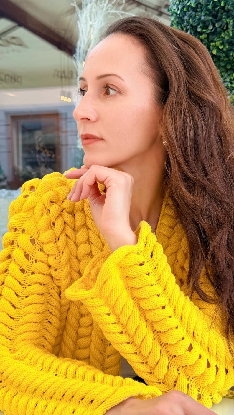 May include: A bright yellow long-sleeved sweater featuring a chunky, braided cable knit pattern with a V-neckline. The garment is constructed with thick, textured yarn, showcasing a prominent vertical weave design throughout the sleeves and bodice.