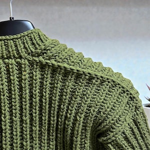 May include: Close-up of a green knitted sweater with a ribbed texture. The sweater is hanging on a black hanger. The olive green color is consistent throughout the garment, highlighting the detailed knit pattern.