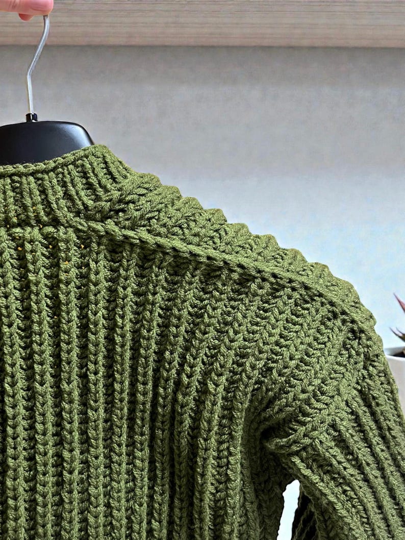May include: Close-up of a green knitted sweater with a ribbed texture. The sweater is hanging on a black hanger. The olive green color is consistent throughout the garment, highlighting the detailed knit pattern.