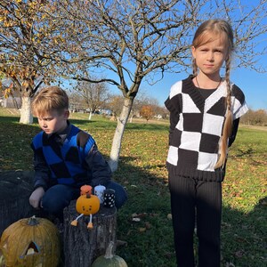 May include: Two children in a sunny outdoor setting. One child wears a black and white checkerboard sweater and black pants. The other child wears a blue and black checkerboard sweater. Pumpkins and candles are nearby.