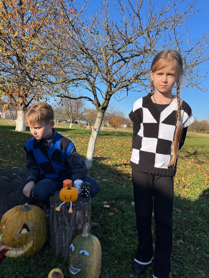 May include: Two children in a sunny outdoor setting. One child wears a black and white checkerboard sweater and black pants. The other child wears a blue and black checkerboard sweater. Pumpkins and candles are nearby.