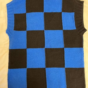 May include: A sleeveless sweater vest in a checkerboard pattern of black and blue squares. The vest has a ribbed hem and neckline. The fabric appears to be a knitted material. The vest is displayed on a light-colored surface.