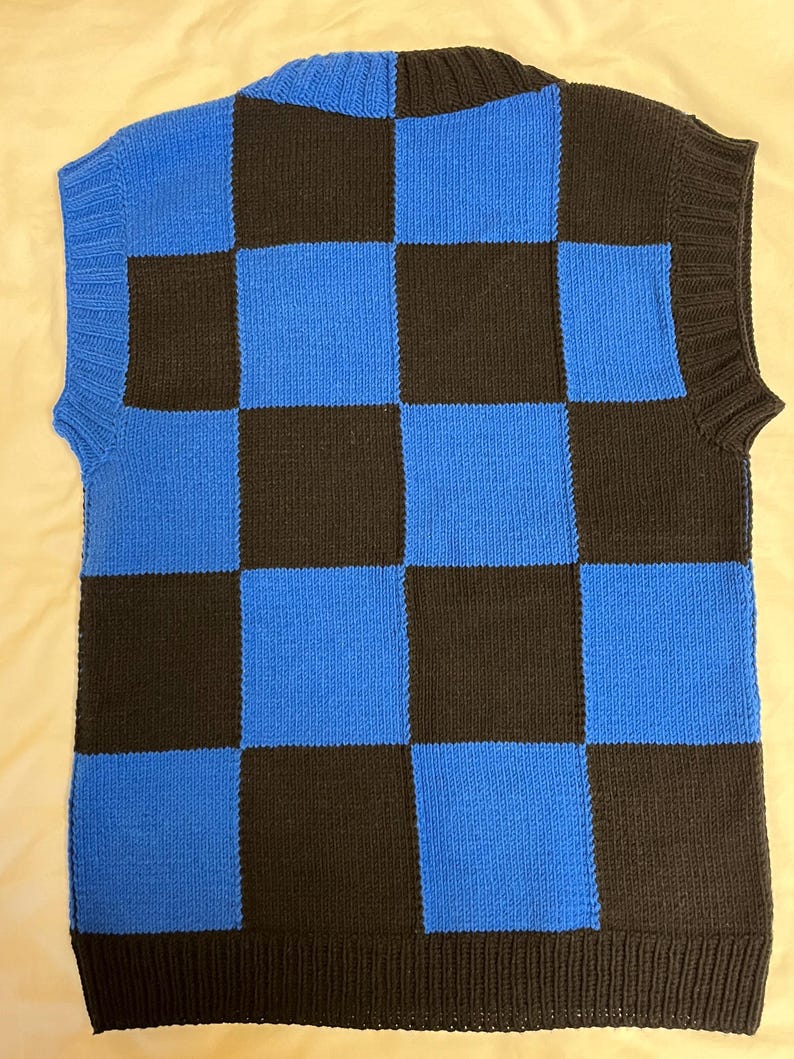 May include: A sleeveless sweater vest in a checkerboard pattern of black and blue squares. The vest has a ribbed hem and neckline. The fabric appears to be a knitted material. The vest is displayed on a light-colored surface.