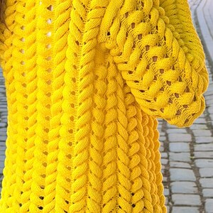 May include: A bright yellow long-sleeved cardigan featuring a prominent vertical cable knit pattern throughout the garment. The knit texture is thick and raised, creating a series of repeating braided columns that extend from the shoulders down.