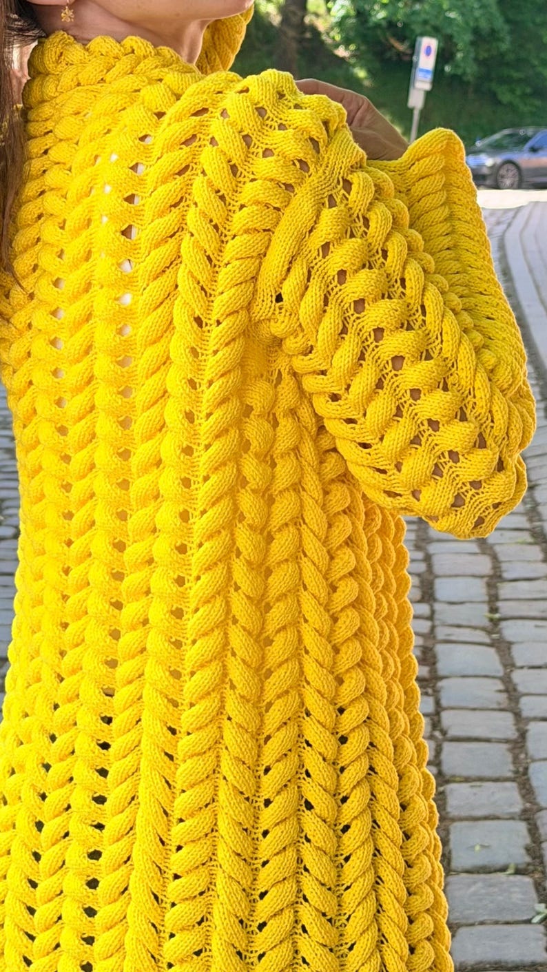 May include: A bright yellow long-sleeved cardigan featuring a prominent vertical cable knit pattern throughout the garment. The knit texture is thick and raised, creating a series of repeating braided columns that extend from the shoulders down.