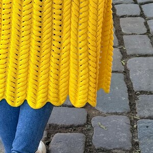 May include: A bright yellow knitted cardigan featuring a vertical braided cable stitch pattern with a scalloped hemline, worn over blue denim jeans and white athletic sneakers. The garment displays a consistent texture and vibrant color throughout.