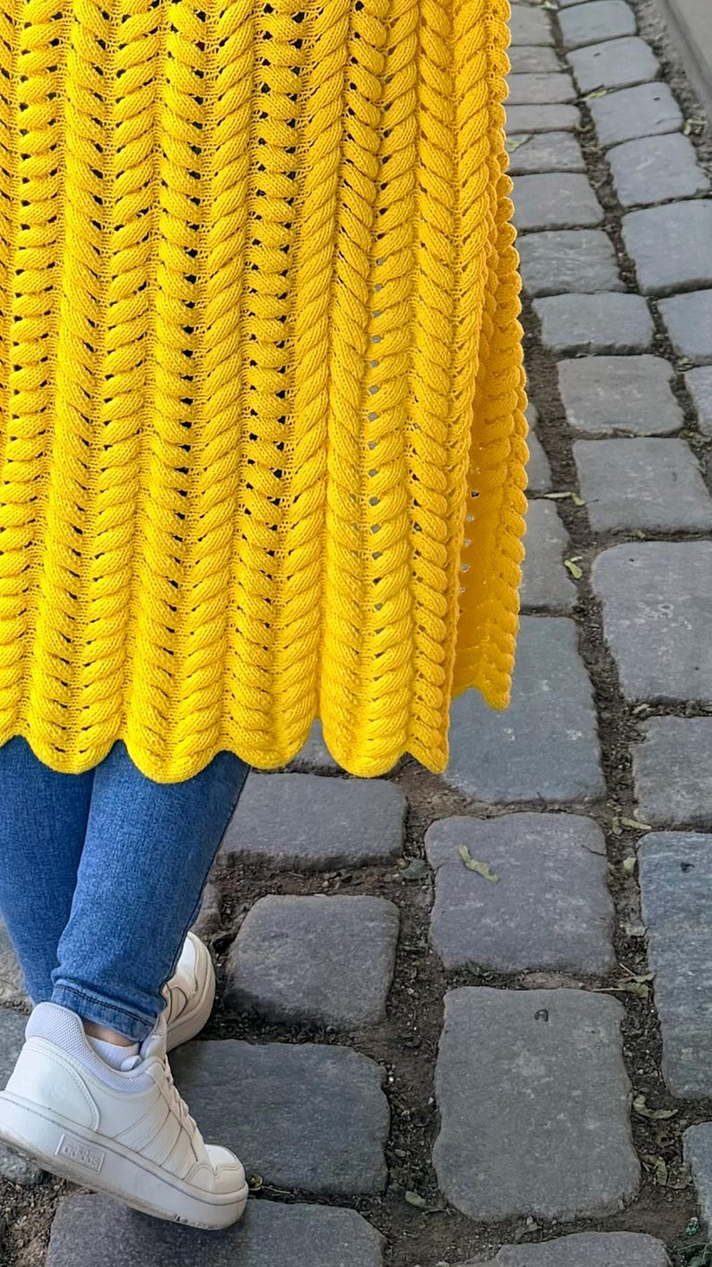 May include: A bright yellow knitted cardigan featuring a vertical braided cable stitch pattern with a scalloped hemline, worn over blue denim jeans and white athletic sneakers. The garment displays a consistent texture and vibrant color throughout.
