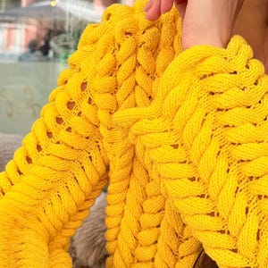 May include: A bright yellow long-sleeve sweater featuring a prominent chunky cable knit pattern with vertical braided textures. The garment has a thick, textured weave throughout the sleeves and body, showcasing a vibrant, solid yellow color.